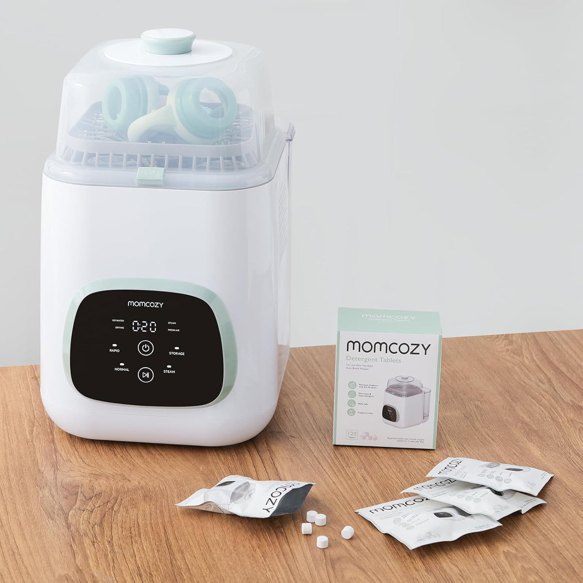 White baby bottle sterilizer with digital display on a wooden surface, accompanied by Momcozy packaging and packets.